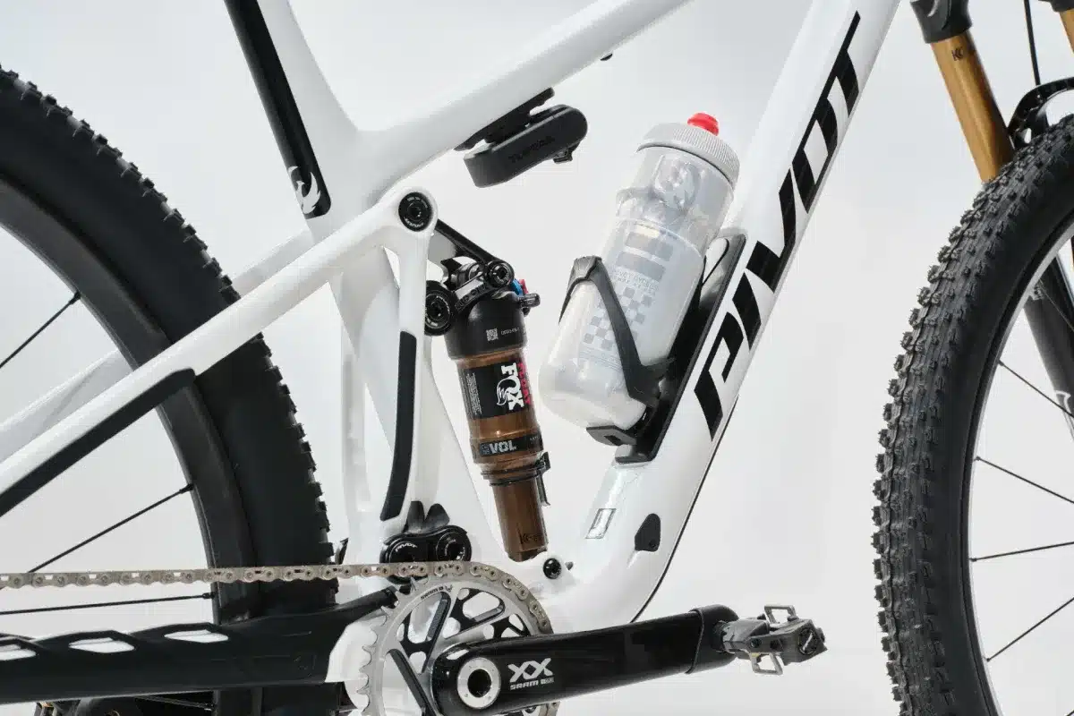 Nahaufnahme des weißen Rahmens eines Pivot Mach 4 SL Mountainbikes, die den hinteren Dämpfer, die Kettenstrebe und die RockShox-Federung zeigt, daneben eine Trinkflasche im Halter.