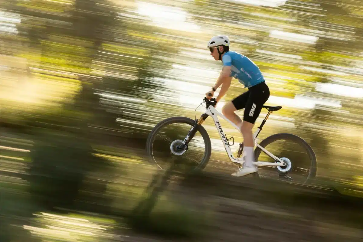 Ein Radfahrer fährt ein weißes Pivot Mach 4 SL Mountainbike auf einem schnellen Trail, umgeben von verschwommenen Bäumen und Sonnenlicht.