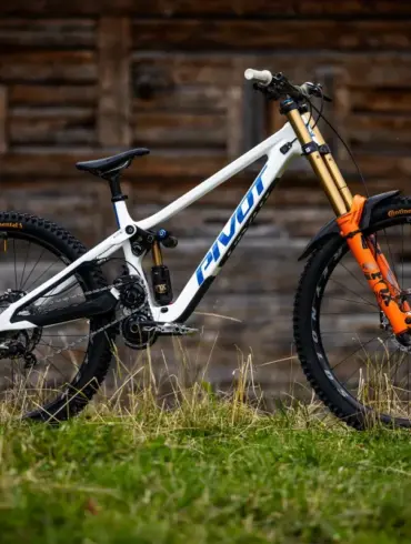 Seitenansicht eines weißen Pivot Phoenix Downhill-Mountainbikes mit orangefarbener Federgabel und schwarzen Laufrädern, stehend im Gras vor einer Holzwand.