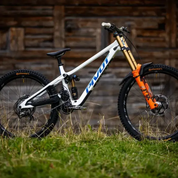 Seitenansicht eines weißen Pivot Phoenix Downhill-Mountainbikes mit orangefarbener Federgabel und schwarzen Laufrädern, stehend im Gras vor einer Holzwand.