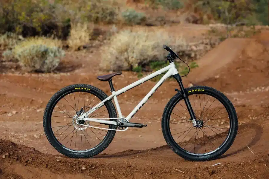 Ein weißes Pivot Point Dirtjump Bike steht auf dem Boden eines Dirtparks, umgeben von Hügeln und Vegetation.