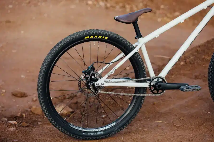 Nahaufnahme des Hinterrads eines weißen Pivot Point Dirtjump Bikes mit Maxxis-Reifen, Scheibenbremse und Singlespeed-Antrieb, auf sandigem Untergrund.