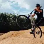 Fahrer mit Helm auf einem Hardtail-Dirtjump-Bike mit 26-Zoll-Rädern und 100 mm Federgabel, der einen Sprung auf einem sandigen Pumptrack ausführt.
