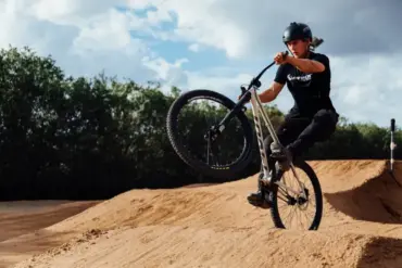 Fahrer mit Helm auf einem Hardtail-Dirtjump-Bike mit 26-Zoll-Rädern und 100 mm Federgabel, der einen Sprung auf einem sandigen Pumptrack ausführt.