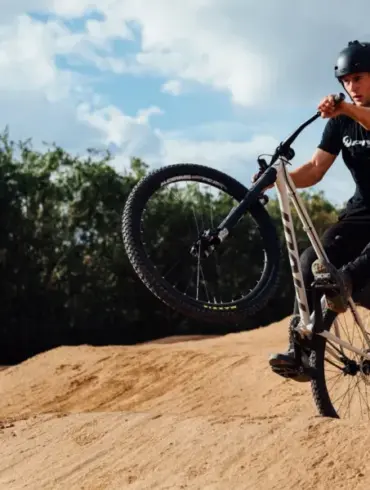 Fahrer mit Helm auf einem Hardtail-Dirtjump-Bike mit 26-Zoll-Rädern und 100 mm Federgabel, der einen Sprung auf einem sandigen Pumptrack ausführt.