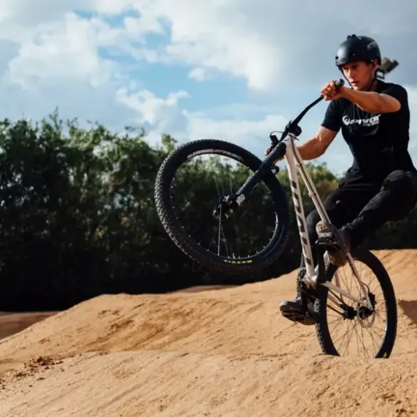 Fahrer mit Helm auf einem Hardtail-Dirtjump-Bike mit 26-Zoll-Rädern und 100 mm Federgabel, der einen Sprung auf einem sandigen Pumptrack ausführt.