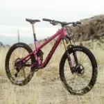 Ein pinkfarbenes Pivot Shadowcat Mountainbike mit 27,5-Zoll-Laufrädern, Fox-Federung und Maxxis-Reifen, das auf einem staubigen Untergrund steht.