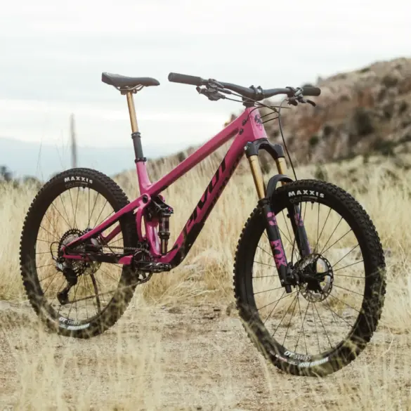 Ein pinkfarbenes Pivot Shadowcat Mountainbike mit 27,5-Zoll-Laufrädern, Fox-Federung und Maxxis-Reifen, das auf einem staubigen Untergrund steht.