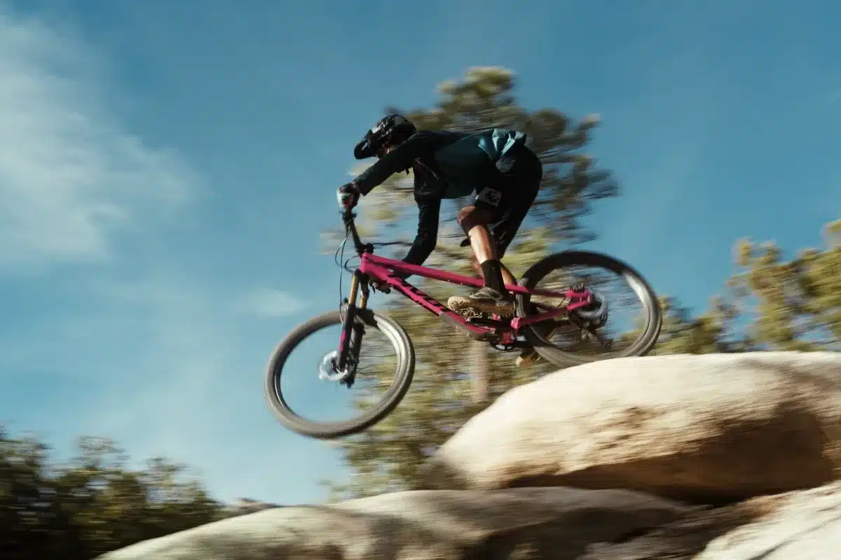 Eine Frau auf einem pinken Mountainbike, dem Pivot Shadowcat, macht einen Sprung über einen Felsen auf einem natürlichen Trail.