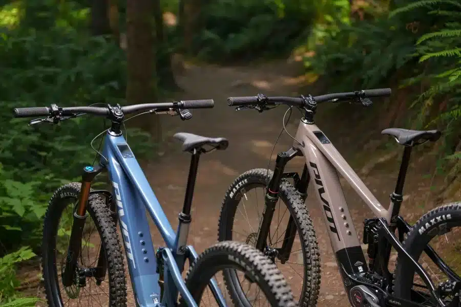 Zwei Pivot Shuttle AM E-Mountainbikes in unterschiedlichen Farben auf einem Waldpfad mit Bäumen im Hintergrund.