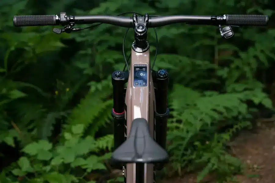 Detailansicht des Cockpits eines braunen Pivot Shuttle AM E-Mountainbikes mit schwarzen Griffen, Display und Federgabel vor einem Hintergrund aus grünem Laub.