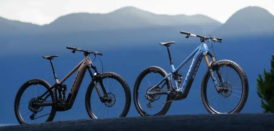 Zwei Pivot Shuttle AM E-Mountainbikes, eines in Badlands Brown und eines in Blue Dark Sky, auf einem Hügel vor bergiger Kulisse.
