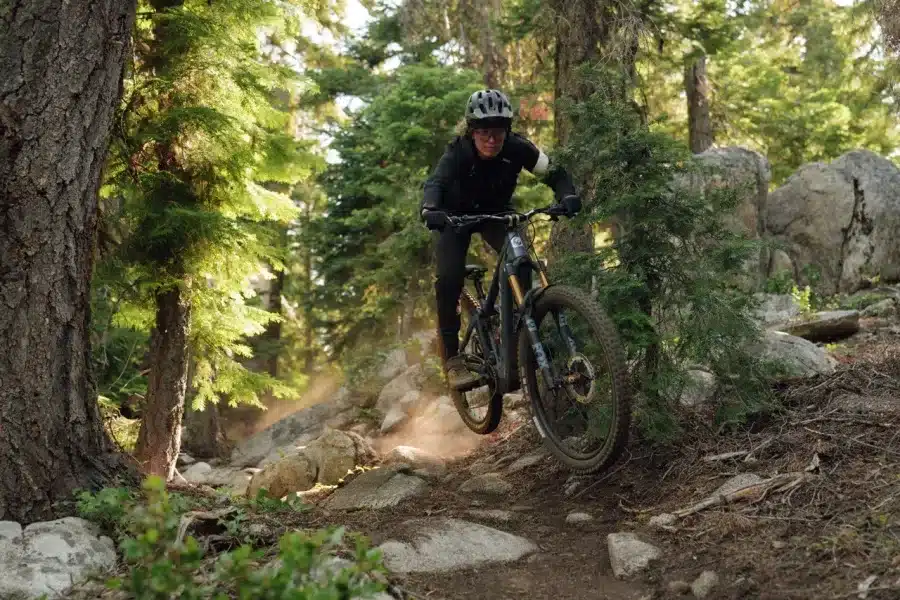 Ein Mountainbiker mit Helm und Protektoren fährt mit einem E-Bike über einen steinigen Pfad in einem Wald und springt über Hindernisse, Staub wirbelt auf.