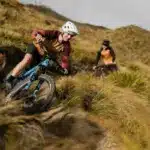 Eine Frau fährt mit einem blau-schwarzen E-Enduro-Mountainbike eine technisch anspruchsvolle Abfahrt auf einem grasbewachsenen Berghang hinunter, während im Hintergrund ein weiterer Fahrer zu sehen ist.