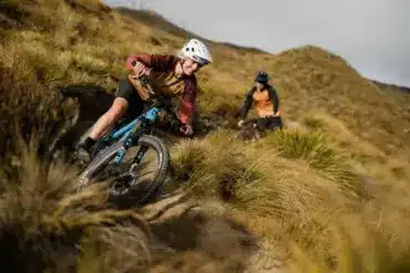 Eine Frau fährt mit einem blau-schwarzen E-Enduro-Mountainbike eine technisch anspruchsvolle Abfahrt auf einem grasbewachsenen Berghang hinunter, während im Hintergrund ein weiterer Fahrer zu sehen ist.
