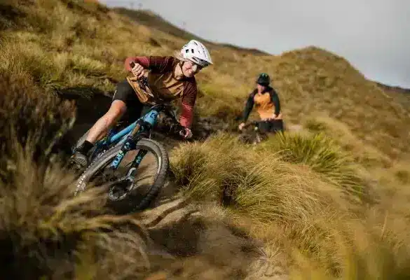 Eine Frau fährt mit einem blau-schwarzen E-Enduro-Mountainbike eine technisch anspruchsvolle Abfahrt auf einem grasbewachsenen Berghang hinunter, während im Hintergrund ein weiterer Fahrer zu sehen ist.