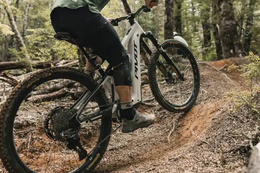 Seitenansicht eines weißen Pivot Shuttle LT E-MTBs, das von einem Fahrer auf einem schmutzigen Waldweg gefahren wird. Der Fokus liegt auf dem Hinterrad und dem Unterrohr des Fahrrads mit dem