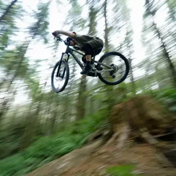 Ein Mountainbiker mit dunklem Trikot und Shorts, der mit seinem Fahrrad in der Luft ist, während er eine Abfahrt in einem Wald fährt.