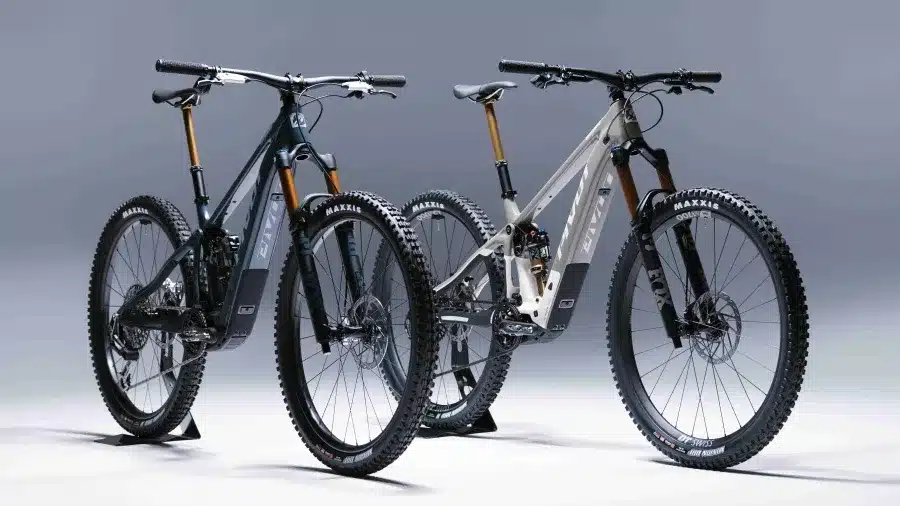 Zwei Pivot Shuttle SL/AM E-Mountainbikes nebeneinander, eines in dunklem Braunton und das andere in glänzendem Grau, beide auf einem hellgrauen Hintergrund.