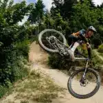Mountainbiker springt mit einem Pivot Switchblade Fahrrad über eine Schanze, umgeben von grüner Natur und einem Waldweg.