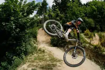 Mountainbiker springt mit einem Pivot Switchblade Fahrrad über eine Schanze, umgeben von grüner Natur und einem Waldweg.