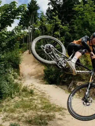 Mountainbiker springt mit einem Pivot Switchblade Fahrrad über eine Schanze, umgeben von grüner Natur und einem Waldweg.