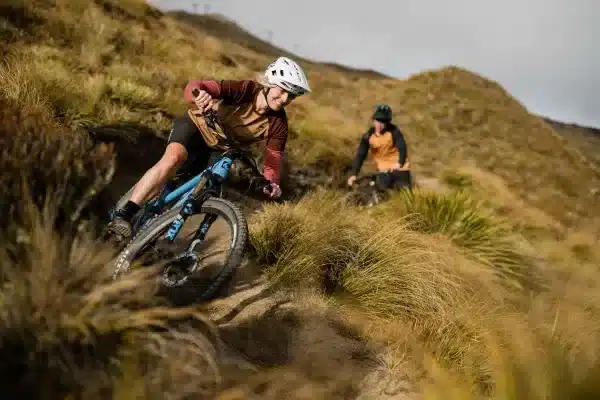 Mountainbikerin in Aktion auf einem schmalen Trail in bergigem Gelände, verfolgt von einer weiteren Person.