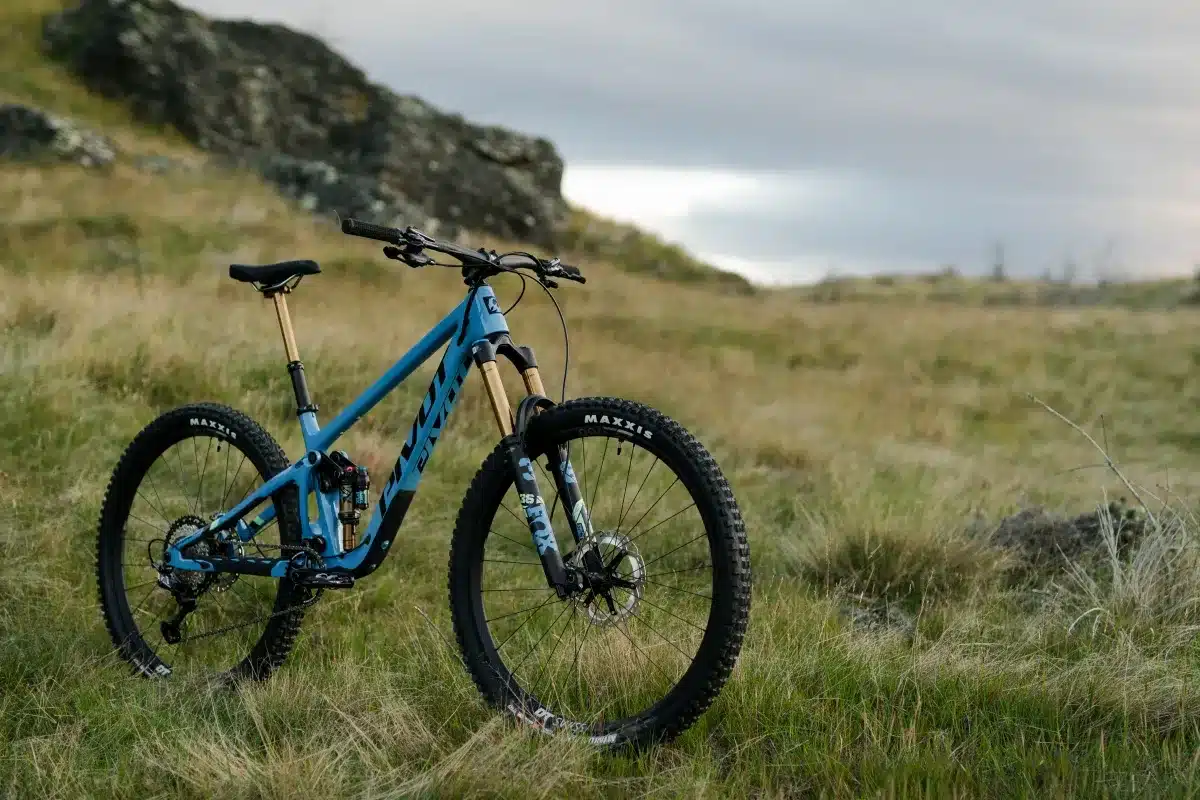Ein blaues Pivot Switchblade Mountainbike mit goldener Sattelstütze und Federgabel, abgebildet im Freien auf einem grasbewachsenen Hügel mit Felsen im Hintergrund.
