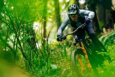 Eine Frau in voller Montur mit Helm und Brille auf einem Mountainbike, das durch einen von Farnen und Bäumen gesäumten Waldweg fährt.