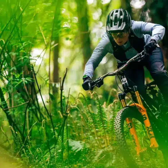 Eine Frau in voller Montur mit Helm und Brille auf einem Mountainbike, das durch einen von Farnen und Bäumen gesäumten Waldweg fährt.