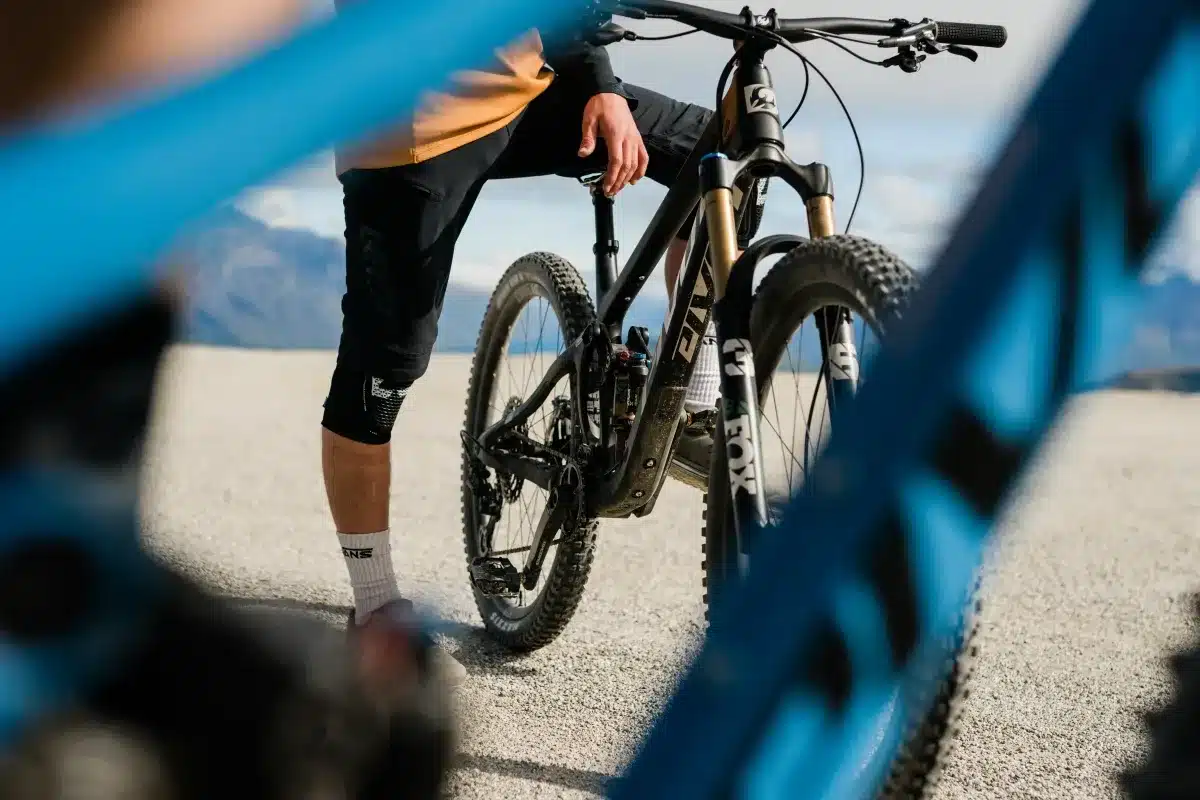 Ein schwarzes Pivot Switchblade Mountainbike mit goldener Fox 36 Gabel, das von einem Fahrer im Freien getragen wird. Ein weiterer blauer Rahmen ist im Vordergrund unscharf.
