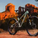 Ein hellgrünes Pivot Trailcat LT Mountainbike steht auf einem roten, staubigen Untergrund vor einer Felsformation im Abendlicht.