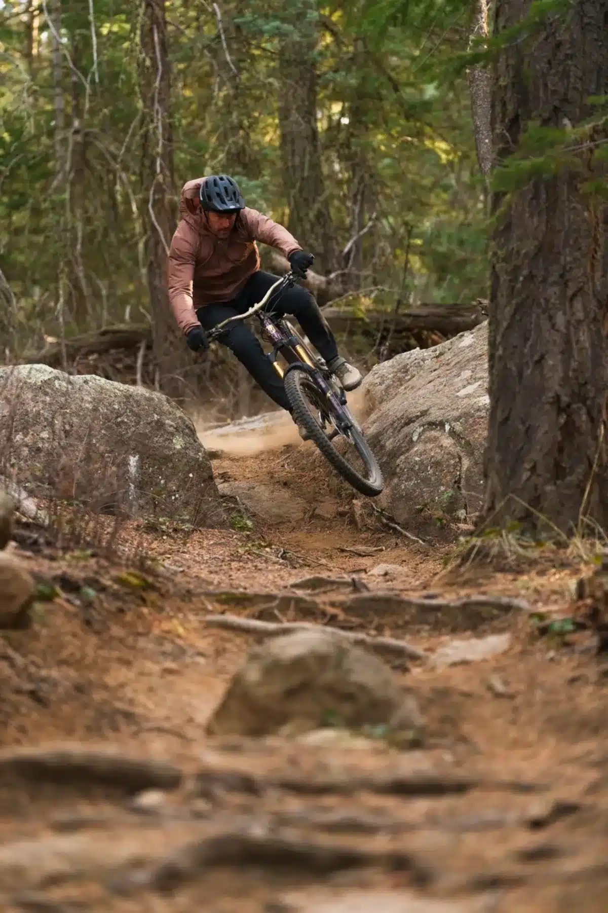 Ein Mountainbiker in brauner Jacke und schwarzer Hose fährt einen felsigen Trail hinunter und erzeugt Staubwolken.
