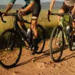 Zwei Gravelbikes, eines in Schwarz und eines in Grün, fahren nebeneinander auf einem staubigen Schotterweg im Freien.