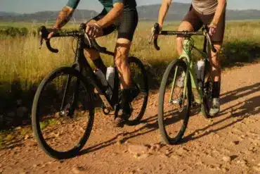Zwei Gravelbikes, eines in Schwarz und eines in Grün, fahren nebeneinander auf einem staubigen Schotterweg im Freien.