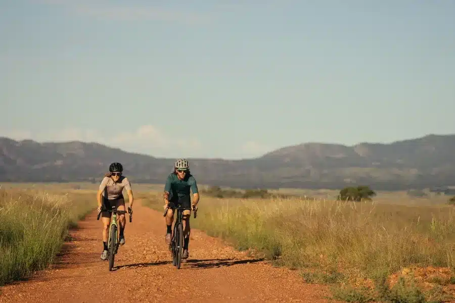 Zwei Radfahrer, ein Mann und eine Frau, fahren mit Gravelbikes auf einem Feldweg in einer weiten, hügeligen Landschaft.