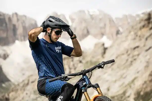 Ein Mann mit Sonnenbrille und Mountainbike-Helm in der Hand, der sich im Freien auf sein Fahrrad setzt.
