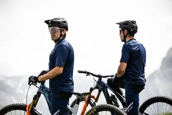 Zwei Mountainbiker stehen nebeneinander mit ihren Rädern. Beide tragen dunkle Mountainbike-Kleidung und schwarze Helme mit Visieren. Der Mann auf der linken Seite blickt nach rechts, während der Mann auf der rechten Seite im Profil nach links schaut.