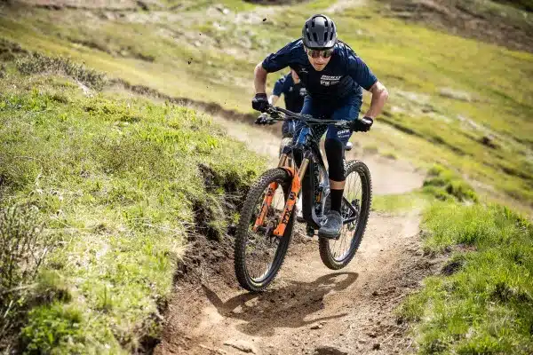 Ein Mountainbiker trägt einen schwarzen Helm und fährt mit einem Full-Suspension-Mountainbike auf einem erdigen Trail bergab, umgeben von grüner Vegetation.
