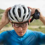 Nahaufnahme eines Rennradfahrers, der seinen silbernen und weißen Rev Pro MIPS Helm aufsetzt, während er Sportbrillen trägt. Hintergrund zeigt eine Landschaft.