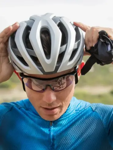 Nahaufnahme eines Rennradfahrers, der seinen silbernen und weißen Rev Pro MIPS Helm aufsetzt, während er Sportbrillen trägt. Hintergrund zeigt eine Landschaft.