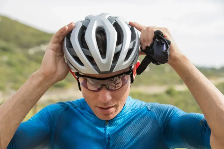 Nahaufnahme eines Rennradfahrers, der seinen silbernen und weißen Rev Pro MIPS Helm aufsetzt, während er Sportbrillen trägt. Hintergrund zeigt eine Landschaft.