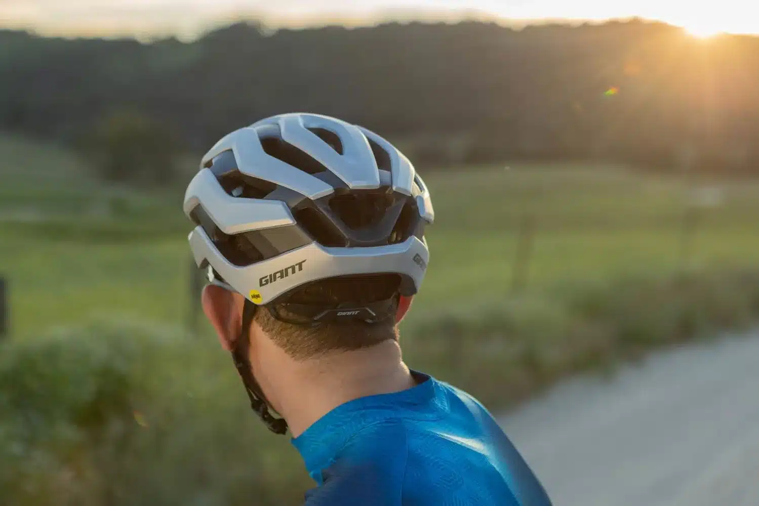 Rückansicht eines silbernen Giant Rev Pro MIPS Fahrradhelms, getragen von einem Mann, der auf einem Feldweg im Sonnenuntergang radelt.