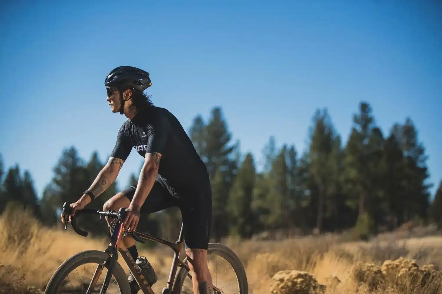 Radsportler mit schwarzem Helm und schwarzer Radsportkleidung fährt auf einem modernen Gravelbike durch eine offene, sonnige Landschaft mit trockenem Gras und wenigen Bäumen unter einem klaren blauen Himmel.