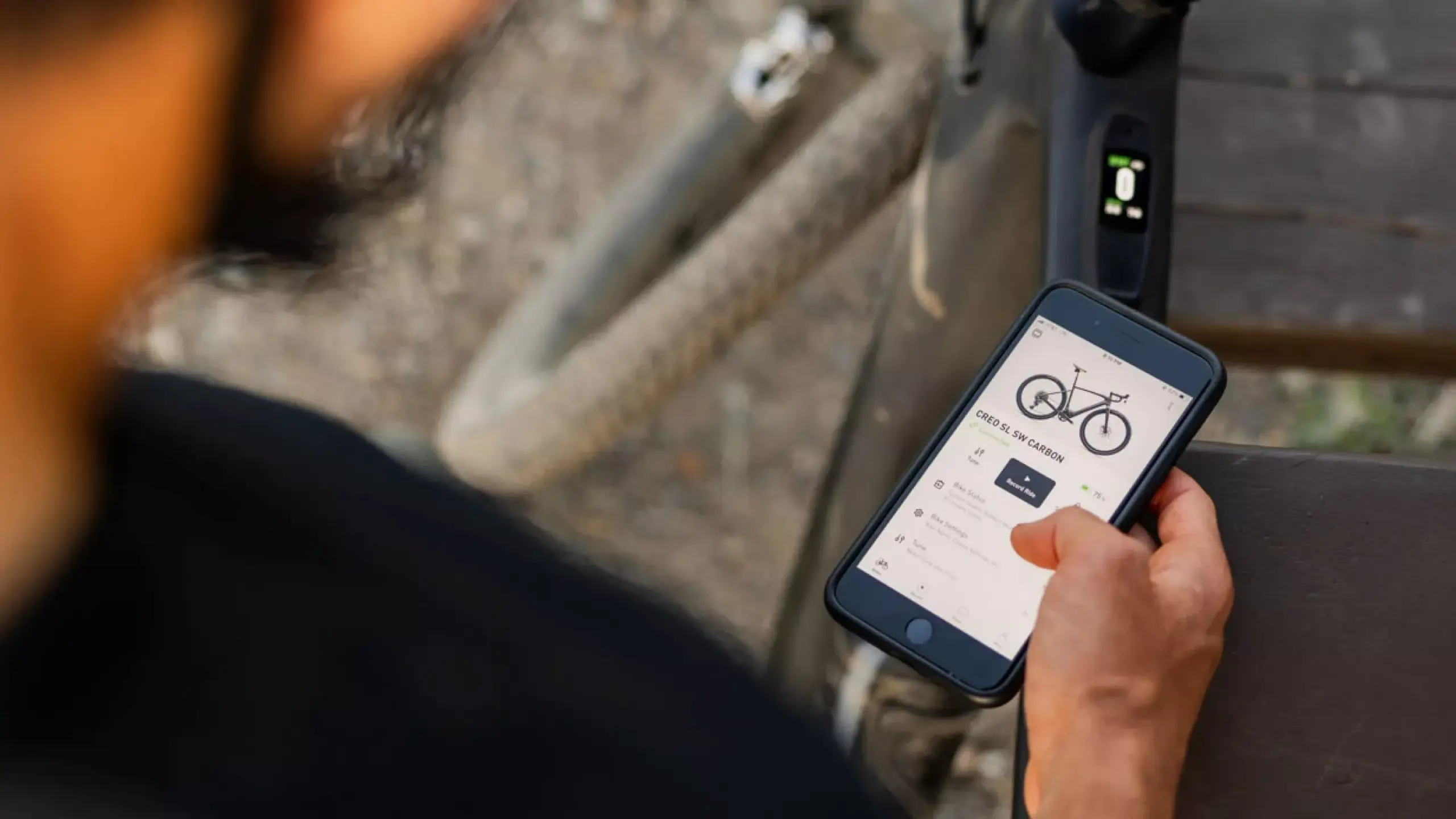 Eine Person hält ein Smartphone in der Hand, auf dessen Bildschirm die Benutzeroberfläche einer Fahrrad-App mit der Abbildung des S-Works Turbo Creo 2 und verschiedenen Einstellungsoptionen sichtbar ist.
