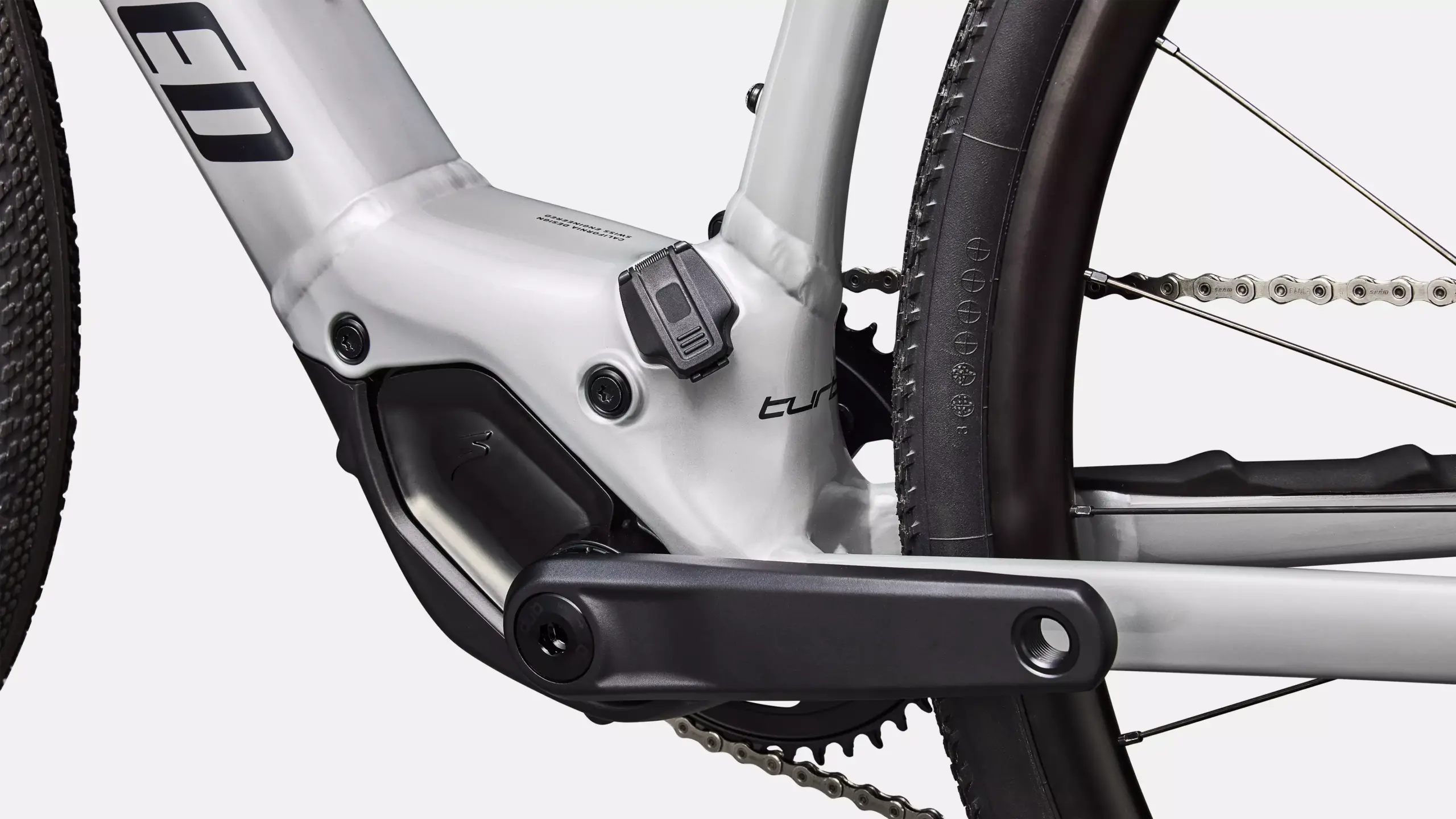 Nahaufnahme des silbernen Rahmens mit dem Schriftzug 'turbo', dem schwarzen Kurbelarm, dem SRAM Eagle Schaltwerk, der Kette und einem Teil des Hinterradreifens eines Specialized Turbo Creo 2 Comp E5 E-Gravelbikes.