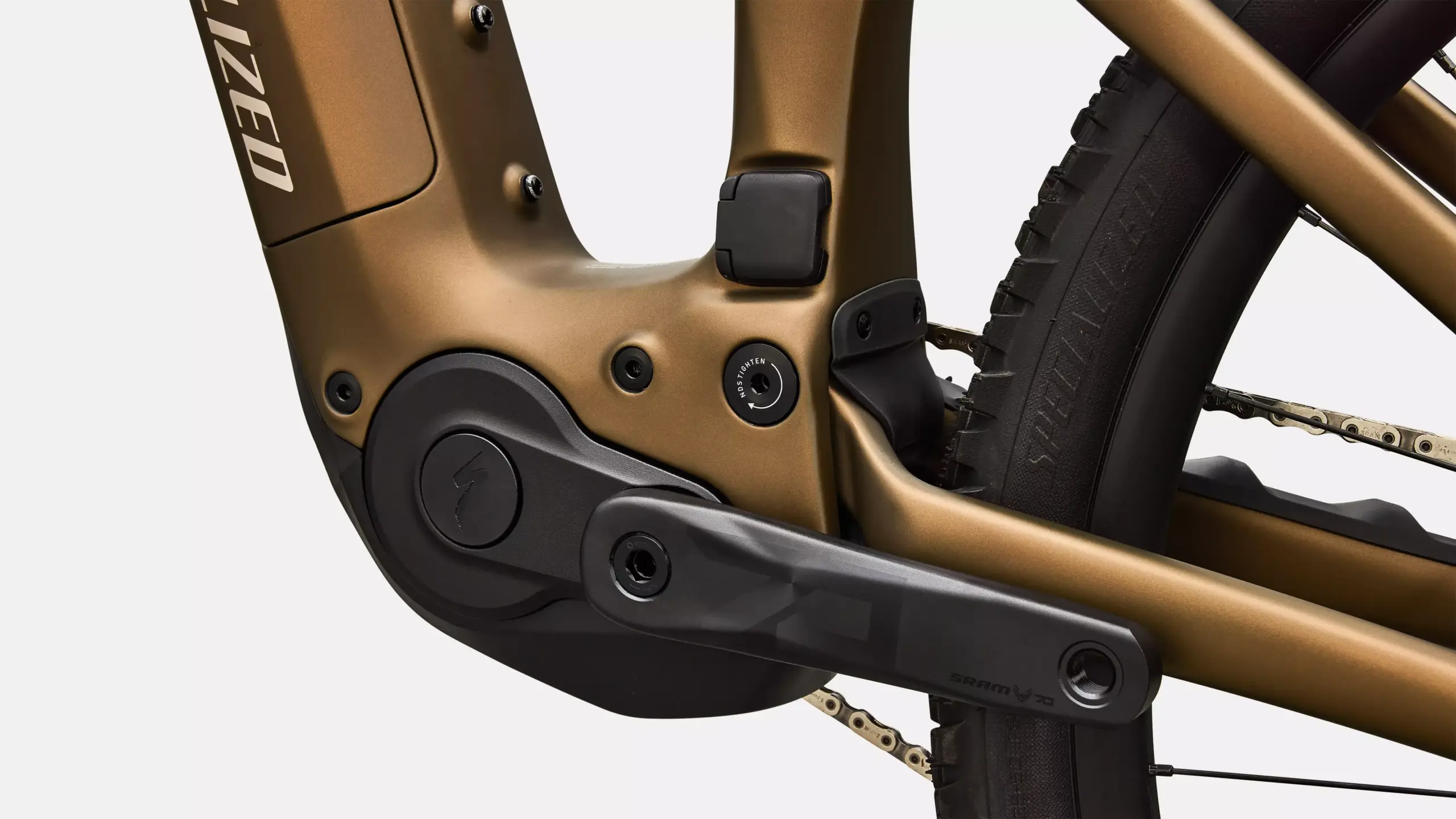 Nahaufnahme des Kurbelarms, der Kette und des Hinterbaus eines Specialized Turbo Levo R Comp E-Mountainbikes in Goldbraun mit schwarzen Akzenten und dem Specialized-Logo.
