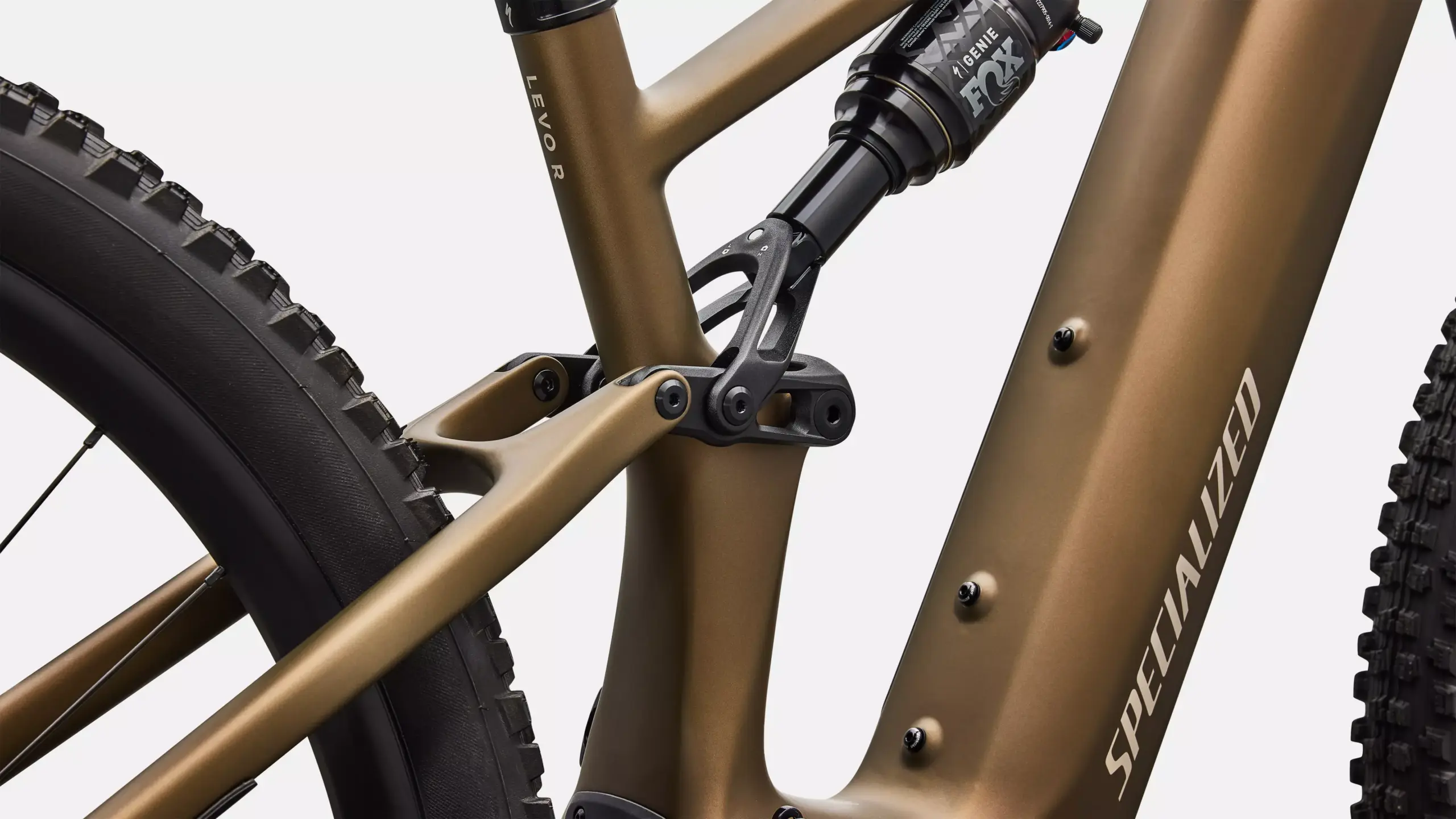 Detailansicht des goldbraunen Rahmens eines Specialized Turbo Levo R Comp E-Mountainbikes, fokussiert auf die Hinterradaufhängung mit Dämpfer und das grobstollige Hinterrad.