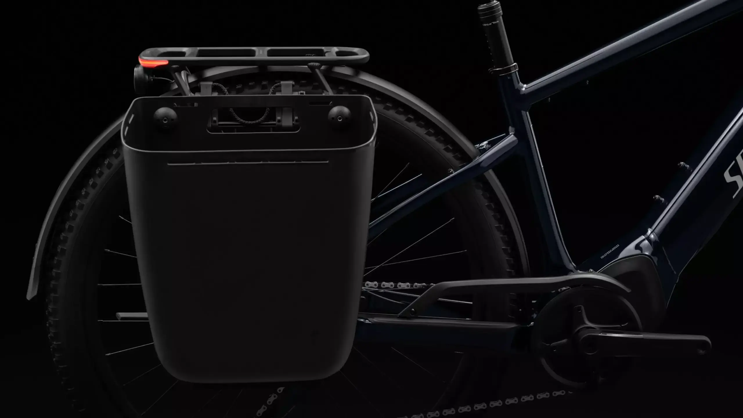 Nahaufnahme des hinteren Teils eines dunkelblauen Specialized Turbo Tero 5.0 EQ E-Bikes, das einen Gepäckträger mit einer abnehmbaren schwarzen Tasche sowie ein integriertes Rücklicht zeigt. Der Hintergrund ist schwarz.