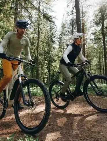 Zwei Personen, ein Mann und eine Frau, fahren nebeneinander auf E-Bikes im Wald auf einem erdigen Weg.